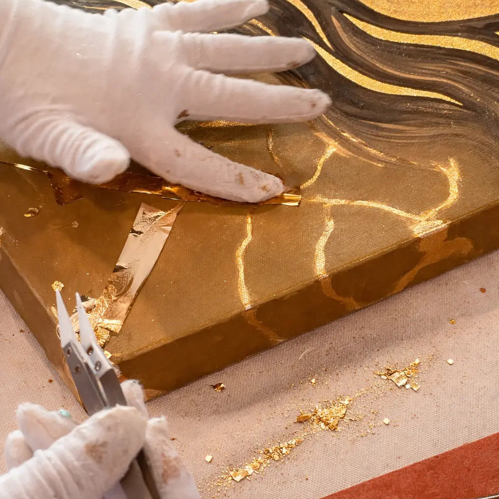 Close-up of an artist applying gold leaf by hand to a painted canvas using tweezers and cotton gloves.